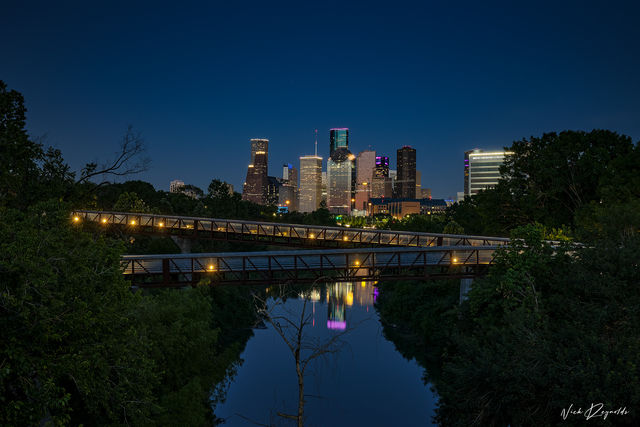 Downtown Houston Texas evening skyline with lit-up pedestrian bridges crossing over the bayou and reflections in the water in the foreground. 