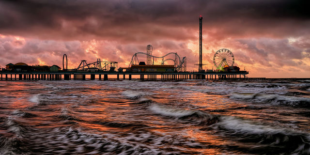 Red and orange overcast sunrise view of the Historic Pleasure Pier in Galveston, Texas with ocean waves in the foreground.